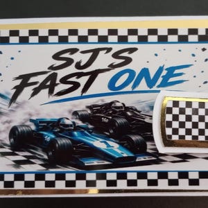 May include: A rectangular card featuring a racing theme with a black and white checkered border and gold trim. The text reads SJ'S FAST ONE above an illustration of a blue race car and a black race car on a track. A small checkered tab with gold edges.