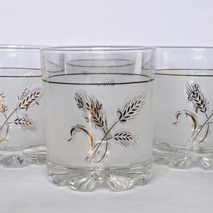 May include: Set of three clear drinking glasses with a frosted wheat design. Each glass has a gold rim and a decorative wheat stalk in gold and black. The glasses have a unique scalloped base.