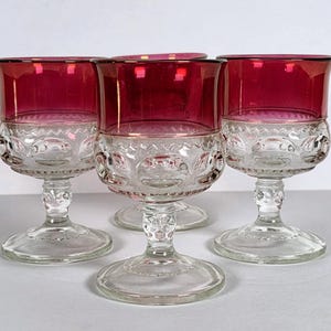 Puede incluir: Cuatro copas de cristal transparente con borde rojo rubí. La parte inferior del cristal presenta un diseño decorativo. Las copas se muestran sobre una superficie blanca. La cristalería es probablemente para servir vino u otras bebidas.