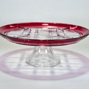 May include: A clear glass serving dish with a ruby red rim and a clear, grid-like interior. The dish sits on a clear, pedestal base. The dish's shadow is visible on the white surface.