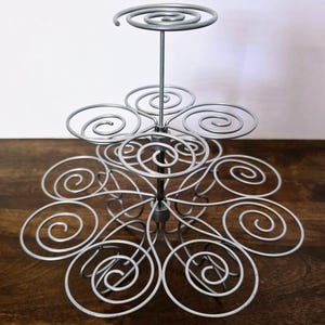 Könnte beinhalten: Ein silberner Cupcake-Ständer aus Metall mit einem Spiraldesign. Der Ständer hat mehrere Ebenen, jede mit einer kreisförmigen Plattform zur Aufnahme von Cupcakes. Die Basis des Ständers hat eine Blumenform.