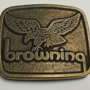 May include: A bronze-coloured belt buckle with an eagle design. The word "browning" is embossed below the eagle. The buckle has a textured surface and a raised border. The buckle is rectangular in shape.