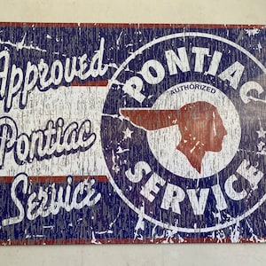 May include: A vintage-style metal sign with a distressed blue and red color scheme. It features the text "Approved Pontiac Service" and a circular logo with the text "Pontiac Authorized Service" and a stylized Native American headdress.
