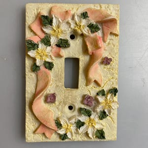 May include: Cream-coloured light switch cover with a raised design. It features white flowers with yellow centres and green leaves, and a pink ribbon. Small purple flowers are also present.