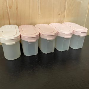 May include: Five clear plastic spice containers with pink lids are arranged in a row. The containers have a rectangular shape with rounded corners. The lids have a flip-top design. The containers are on a dark surface with a light wood background.