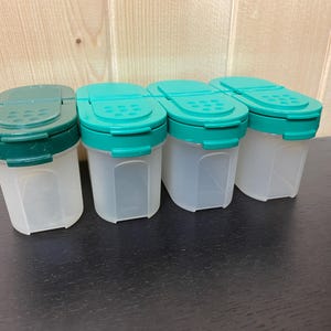 May include: Four clear plastic spice containers with green and teal lids. The lids have a flip-top design with small holes for dispensing. The containers are arranged in a row on a dark surface.
