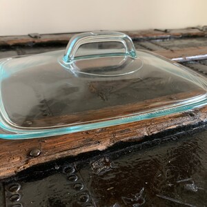May include: A clear, square glass lid with a rectangular handle. The lid has a slightly rounded edge and is designed for cookware. The handle is arched and integrated into the lid's design.