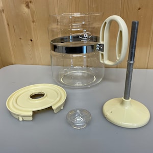 May include: A disassembled clear glass coffee maker with a cream-coloured handle, base, and lid. The glass carafe has a silver band and the brand name "Genco Brand" is visible. The components are arranged on a grey surface.