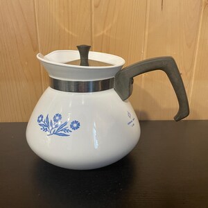May include: White enamel coffee pot with a black handle and lid. The pot features a silver band and blue floral design. The handle is dark grey. The pot is on a dark surface with a light wood background.