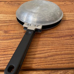 May include: A small, silver-coloured, hinged egg frying pan with a black handle. The pan is round and has a lid. The handle has a hole at the end for hanging. The pan is on a wooden surface.