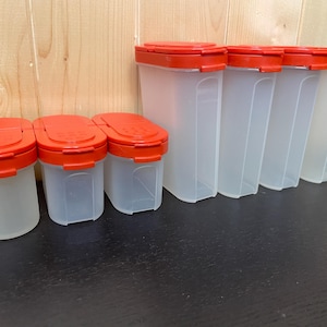 Peut inclure: Ensemble de récipients à épices en plastique transparent avec des couvercles rouges vifs. Les récipients sont rectangulaires et de différentes tailles, avec un total de neuf récipients visibles. Les couvercles sont à rabat pour un accès facile.