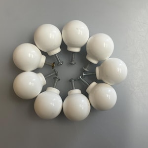 May include: Ten white ceramic cabinet knobs arranged in a circle. Each knob has a rounded, spherical shape and is attached to a metal screw. The knobs are set against a plain, light grey background.