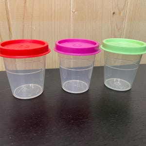 May include: Three clear plastic cups with colored lids. The lids are red, magenta, and green. The cups are on a dark surface with a light wood background.