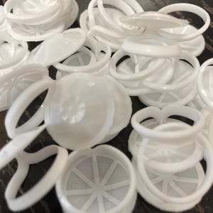 Peut inclure: Gros plan sur des bouchons de bouteilles en plastique blanc et des composants en forme d'anneau. Les bouchons ont un design circulaire avec un motif en étoile à l'intérieur. Les anneaux sont incurvés.