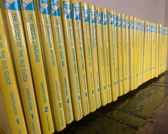 May include: A row of vintage yellow hardback books, including titles such as "The Secret of the Old Clock" and "The Hidden Staircase." The spines have blue detailing and the author's name, Carolyn Keene, with visible book numbers.