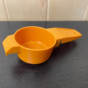 May include: An orange plastic kitchen tool with a round cup and a handle. The cup has a perforated bottom. The handle has a flat, rectangular shape. The tool is designed for straining or measuring ingredients.