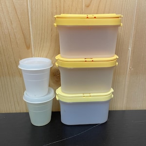 May include: A collection of clear plastic food storage containers with yellow lids and small white cups with lids. The containers are stacked on a dark surface against a light wood background. The containers are designed for food storage.