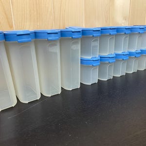 May include: A collection of clear plastic food storage containers with blue lids. The containers are rectangular with rounded corners and are arranged in rows. The lids are a solid blue color. The containers are of varying sizes.