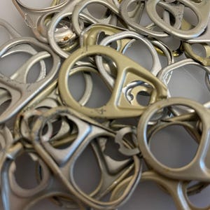 May include: A close-up shot of a pile of silver and gold-colored can pull tabs. The tabs are made of metal and have a circular design with a small tab for lifting. Some tabs show signs of wear.