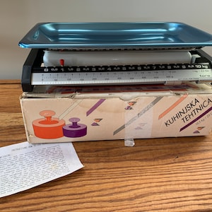 Peut inclure: Balance de cuisine vintage bleue et noire avec un plateau rectangulaire. La balance a un cadran blanc avec des mesures noires et un indicateur rouge. La balance est sur une boîte avec du texte en langue étrangère. Mesure jusqu'à 10 kg.