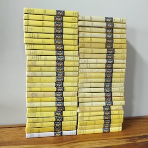 May include: A collection of vintage yellow hardcover books, neatly stacked on a wooden surface. The books have a consistent design with a circular emblem and text on the spine. The spines are yellow with blue and white details.