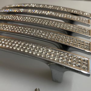 May include: A collection of silver cabinet handles, each embellished with rows of clear, sparkling crystals. The handles have a curved shape and are arranged in a slightly overlapping pattern, highlighting their decorative nature.