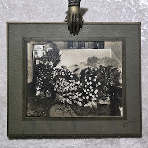 May include: An antique framed photograph featuring a black and white image of floral arrangements. The frame is a muted green with a textured pattern. A decorative bronze hand holds the frame.
