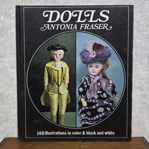 May include: A book titled "DOLLS ANTONIA FRASER" with a black cover. The cover features two antique doll illustrations inside a white circle. The dolls are dressed in period clothing. The text "140 illustrations in color & black and white" is at the bottom.