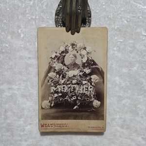 May include: A sepia-toned vintage photograph on a rectangular card featuring a portrait of a person centered within a large floral wreath. The word MOTHER is spelled out in flowers below the portrait. The text WEATHERBY and PLYMOUTH, O. appear.
