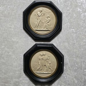 Op de afbeelding: Twee antieke wandplaten met zwarte achthoekige frames. Elke plaat heeft een cirkelvormige, crèmekleurige reliëfsculptuur die klassieke figuren afbeeldt. Eén toont een man die fluit speelt, een hond en een gezin.