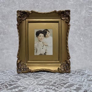 Peut inclure: Un cadre photo rectangulaire doré avec des détails floraux en relief sur les coins. Le cadre contient une photographie de style vintage en sépia représentant deux personnes posant ensemble, l'une debout derrière l'autre en portrait.