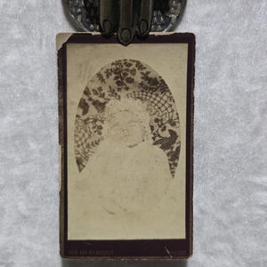 May include: Antique sepia-toned photograph of a person, likely from the Victorian era. The portrait is set against a patterned backdrop within an oval frame. The photograph is held by a decorative metal clip.