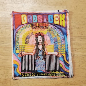 May include: A colorful vintage-style patch with the word "Woodstock" in large, stylized letters. The patch features a woman with long brown hair wearing a flower crown and a blue dress. The text "3 Days of Peace and Music" is at the bottom of the patch.