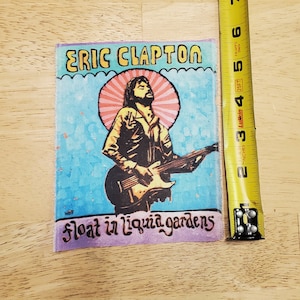 May include: A colorful embroidered patch featuring a cartoon image of Eric Clapton playing a guitar. The patch has a blue background with a pink and orange sun behind Clapton. The text "float in liquid gardens" is written in a purple font at the bottom of the patch.
