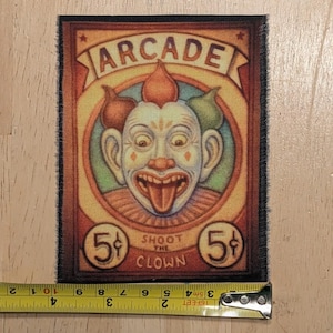 May include: A rectangular fabric patch featuring a vintage arcade game graphic of a clown with a wide grin and tongue out. The text reads ARCADE and SHOOT THE CLOWN with 5 cent symbols. A yellow measuring tape shows the patch is 4 inches wide.