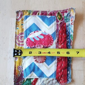 May include: A colourful, handmade fabric patch with various patterns and textures. The patch features a blue and white chevron pattern, a red strawberry shape, and other patterned fabrics. The patch is approximately 10 cm wide.