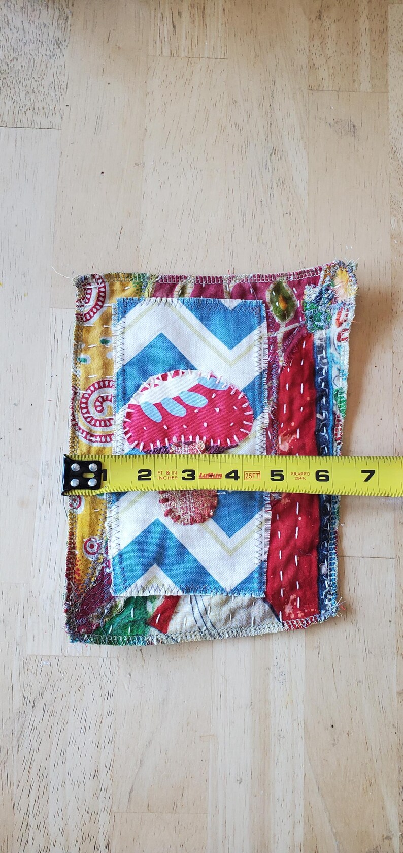 May include: A colourful, handmade fabric patch with various patterns and textures. The patch features a blue and white chevron pattern, a red strawberry shape, and other patterned fabrics. The patch is approximately 10 cm wide.