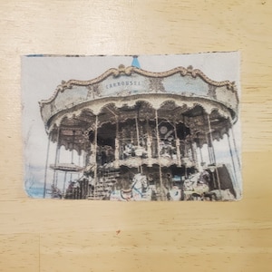May include: A rectangular fabric item featuring a vintage carousel design. The carousel has a light blue and gold color scheme, with the word "CARROUSEL" across the top. The image has a faded, antique aesthetic.