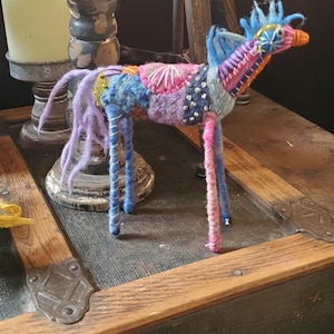 May include: A colourful, whimsical, handmade felt horse sculpture with a blue, pink and purple mane and tail. The horse is standing on a wooden surface.