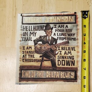 Pu&ograve; includere: Stampa quadrata con un dipinto di Robert Johnson che suona la chitarra. La stampa include il testo che recita "Hellhound on my trail" e "King of the Delta Blues". La stampa &egrave; alta circa 15 cm.