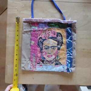 May include: A small, beige tote bag with a blue handle. The bag is made of patchwork fabric and features a colorful image of Frida Kahlo.