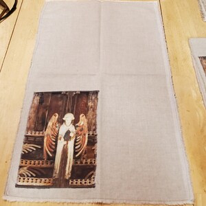 May include: A rectangular linen towel with a printed image of a carved angel figure. The angel has outstretched wings and is standing in front of a wooden door. The towel has a raw edge.