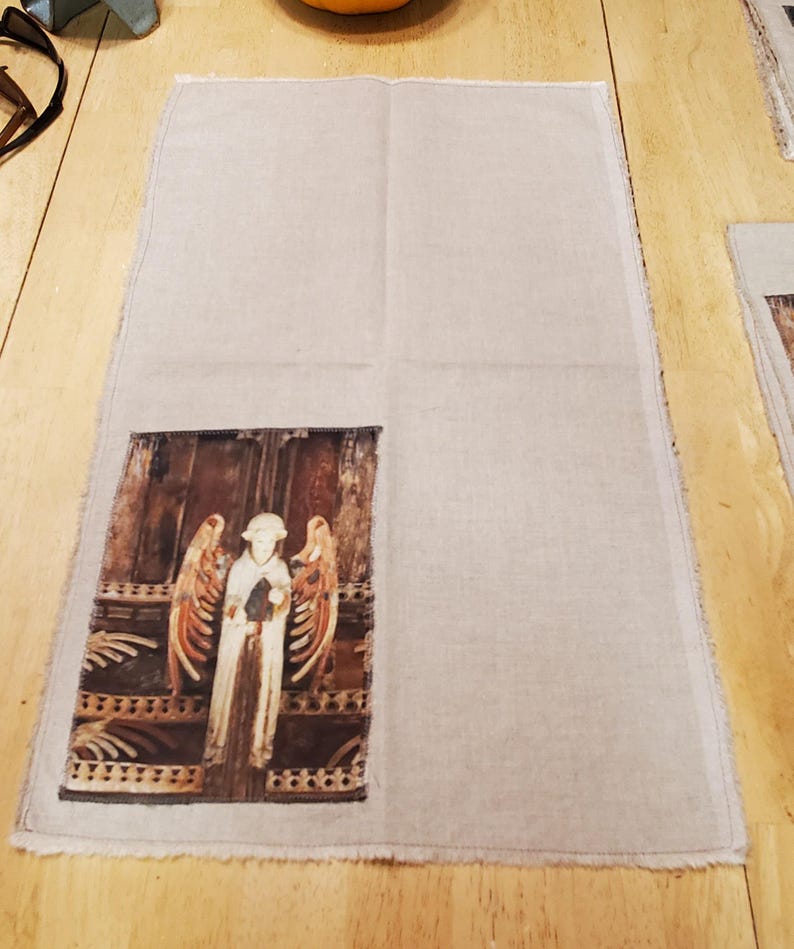 May include: A rectangular linen towel with a printed image of a carved angel figure. The angel has outstretched wings and is standing in front of a wooden door. The towel has a raw edge.