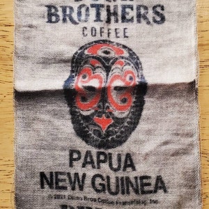 Könnte beinhalten: Ein weißer Stoffbeutel mit schwarzem Text, der "Dunn Brothers Coffee Papua New Guinea" lautet, und einem schwarzen und roten Tribal-Masken-Design. Der Beutel trägt auch den aufgedruckten Text "DB1506".