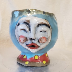 May include: A whimsical ceramic face planter with a blue hood, white face, and red lips. The cheeks are pink, and the base is red with yellow polka dots. The handmade art piece is a unique decorative item.