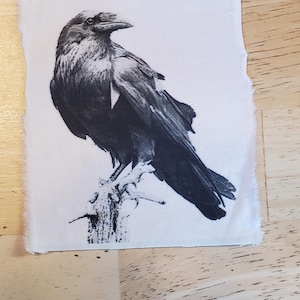 May include: A black and white illustration of a raven perched on a branch. The raven is detailed with its feathers and beak visible. The artwork is printed on a white fabric with a raw edge.