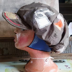 May include: A distressed, olive green newsboy cap with multiple fabric patches in white, orange, and beige. The cap is displayed on a mannequin head with a blue neck and pink cheeks. A silver safety pin is attached to the side.