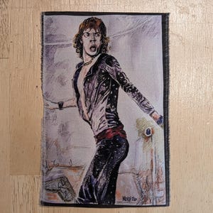 May include: A rectangular fabric patch featuring a detailed illustration of a person with shoulder-length brown hair, wearing a dark open jacket and dark trousers. The figure is captured in a dynamic pose with an open mouth. The signature Wolf 26 is visible in the bottom right corner.