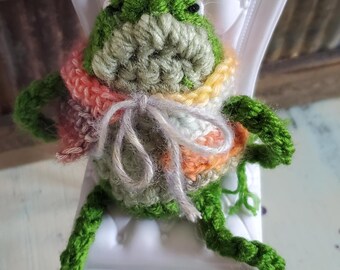 Può includere: Una bambola a forma di rana all'uncinetto fatta a mano con un corpo verde e una sciarpa colorata, seduta su una piccola sedia bianca. La rana ha occhi neri ed è fatta di filato. La sedia ha uno schienale decorativo.