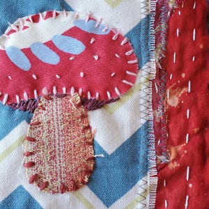 May include: A textile artwork with a red mushroom appliqu&eacute;, white stitching, and a blue and white chevron pattern. The mushroom has a woven stem. Other fabric patterns form the background.